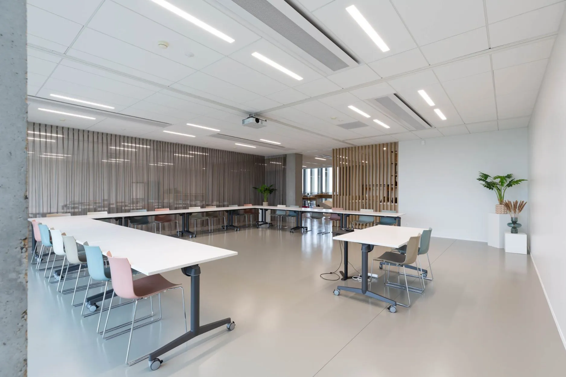 Lichte vergaderzaal met witte tafels in U-opstelling, gekleurde stoelen, plafondbeamer en houten lamellen en gordijnen als scheidingswand