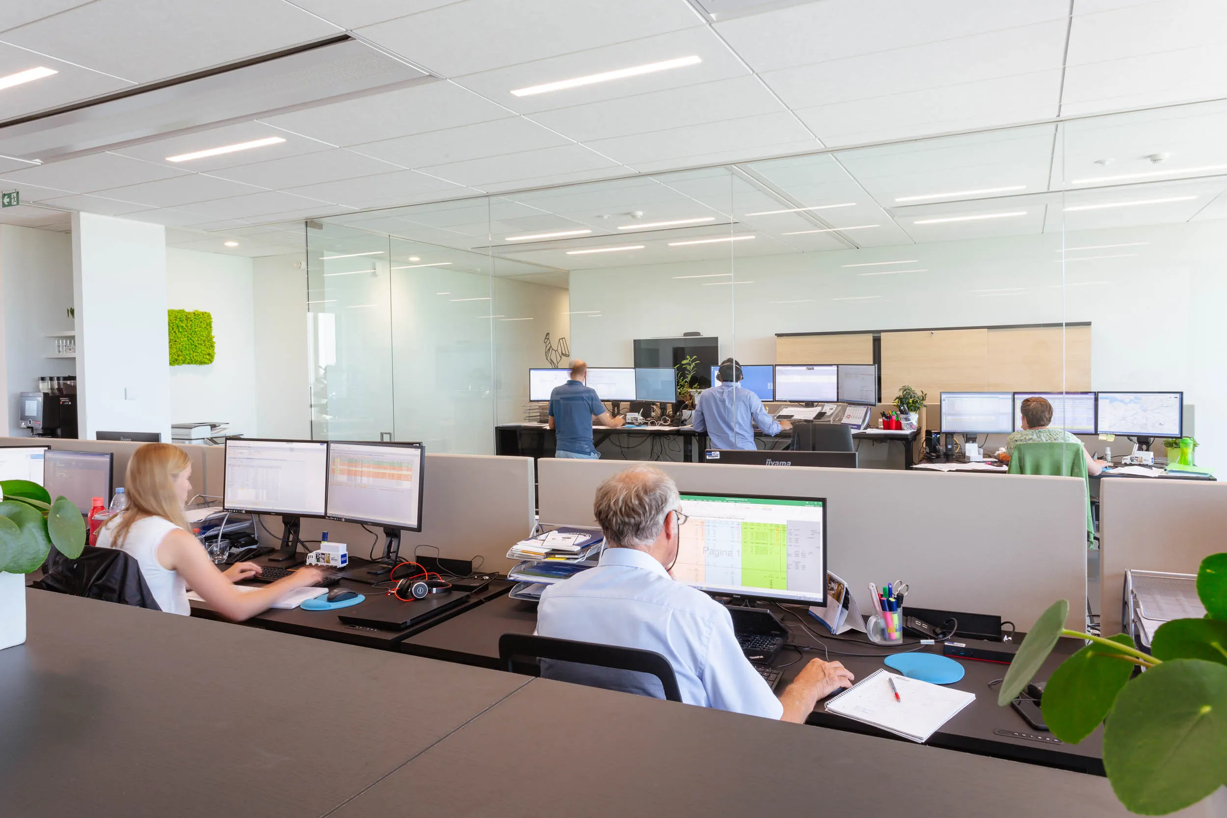 Medewerkers werken achter meerdere schermen in een lichte, moderne open kantoorruimte met glazen wanden en planten.