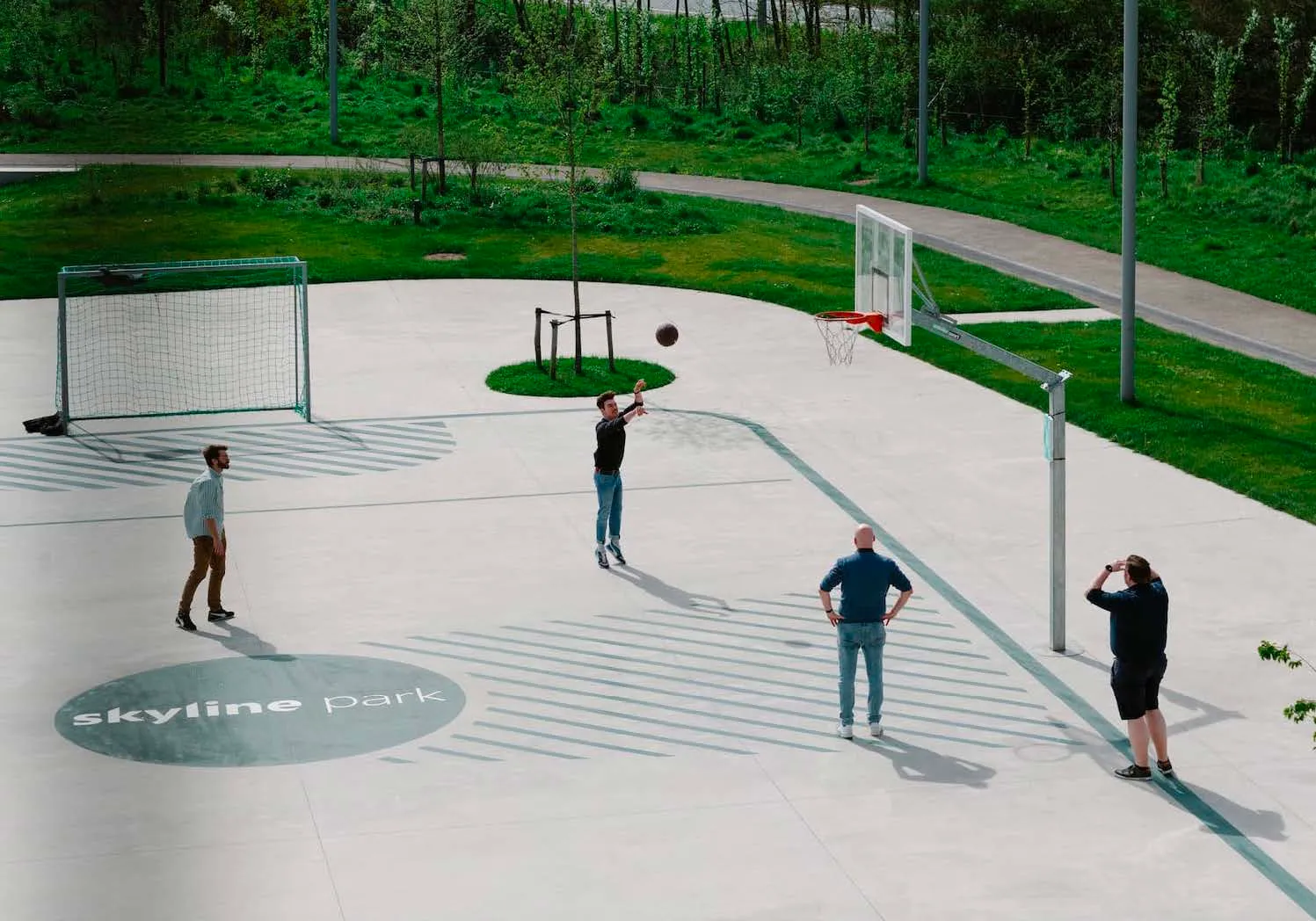 Mensen basketballen op buitensportplein van Skyline Park, omringd door groen, met voetbaldoel en basketbalring.