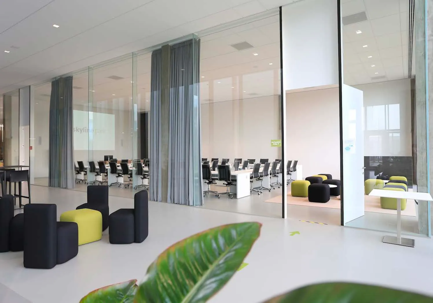 Lichtrijke trainingsruimte met glazen wanden, zwarte bureaustoelen, groene en zwarte poufjes en een projectiescherm op de achtergrond.